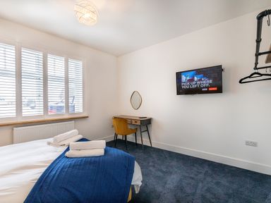 A bedroom with a bed with towels on it a wooden desk with a yellow chair a wall-mounted television and a window with white blinds at Groves House in Castletown near Fortuneswell