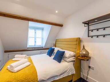 A bedroom with a yellow bed headboard white bed linens blue pillows folded white towels a window with blinds and a wall-mounted lamp at Groves House in Castletown near Fortuneswell