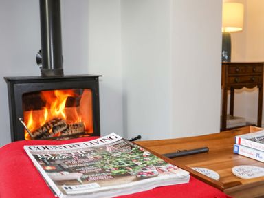 A wood burning stove with fire burning a magazine on a red surface and a wooden table with books and coasters at Cwrt in Dwyran near Niwbwrch Newborough