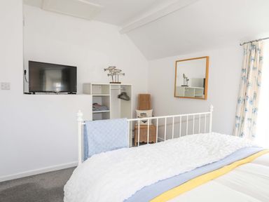 A bedroom with a bed a television on a white shelf an open closet with hangers a wicker chair a mirror on the wall and floral curtains at Cwrt in Dwyran near Niwbwrch Newborough