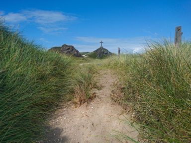 A sandy path through tall grass leading to a cross on a hill under a blue sky at Cwrt in Dwyran near Niwbwrch Newborough