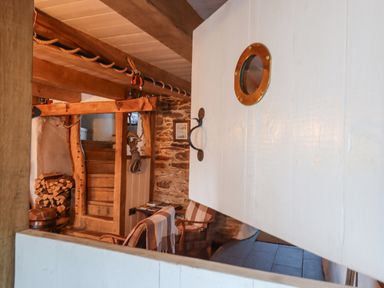 A rustic interior with wooden beams a wooden staircase firewood stacked two chairs with plaid throws and a white half door with a porthole window at Alpha Cottage in Dittisham near Dartmouth