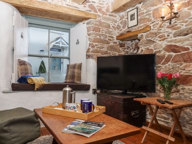 A rustic living room with a window seat, a television on a chest, a wooden table with mugs and a teapot, and a smaller table with flowers at Alpha Cottage in Dittisham near Dartmouth