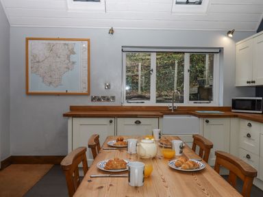 A kitchen with a wooden table set for breakfast croissants orange juice mugs a teapot wooden chairs a window a map on the wall and white cabinets at Alpha Cottage in Dittisham near Dartmouth