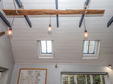 A ceiling with exposed beams hanging light bulbs and two small skylight windows above a wall with a map and a window with a roller blind at Alpha Cottage in Dittisham near Dartmouth