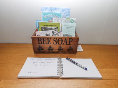 A wooden box labeled BEE SOAP holding brochures and booklets on a wooden surface with an open spiral notebook and pen in front at Alpha Cottage in Dittisham near Dartmouth