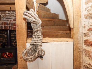 A wooden staircase with a coiled rope hanging from the banister near a stone wall with a lit wood stove at Alpha Cottage in Dittisham near Dartmouth