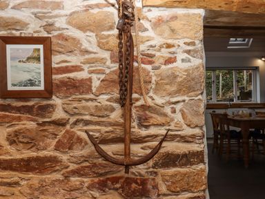 A stone wall with a hanging old rusted anchor and a framed painting next to a kitchen with a wooden dining table and chairs at Alpha Cottage in Dittisham near Dartmouth