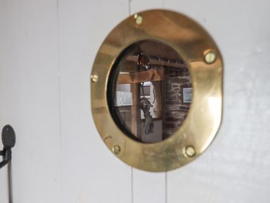 A wooden and stone interior reflected through a round brass porthole window at Alpha Cottage in Dittisham near Dartmouth