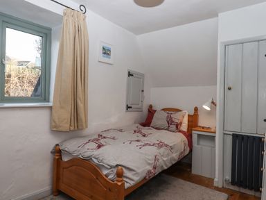 A small bedroom with a single wooden bed a bedside table with a lamp a window with beige curtains and a framed picture on the wall at Alpha Cottage in Dittisham near Dartmouth