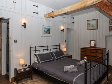 A bedroom with a black metal bed with dark bedding two bedside tables with lamps a wooden dresser and framed pictures on white paneled walls at Alpha Cottage in Dittisham near Dartmouth