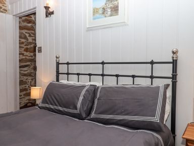A bed with dark gray bedding and pillows against a white paneled wall with a small lamp and an open door showing a stone wall at Alpha Cottage Dittisham near Dartmouth