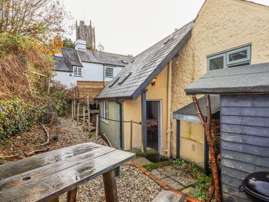 An outdoor area with a gravel path wooden table small shed and a light on a pale yellow house at Alpha Cottage in Dittisham near Dartmouth