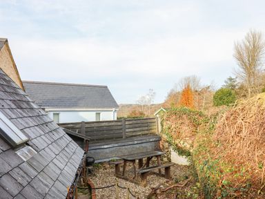 An outdoor patio area with a wooden picnic table and benches gravel ground wooden fencing and surrounding vegetation at Alpha Cottage in Dittisham near Dartmouth