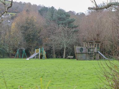 A playground with swings slides and a wooden play structure on a grassy field with trees in the background at Alpha Cottage in Dittisham near Dartmouth