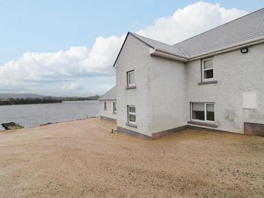A gravel area next to a white house by a body of water with cloudy sky at Dock at Dominic's Ballyheerin near Downings