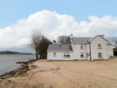 A white house with a gray roof near a body of water and trees at Dock at Dominic's in Ballyheerin near Downings