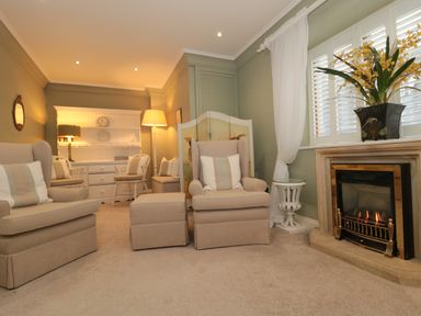 A living room with beige armchairs and ottomans a fireplace with flowers on the mantel and a white cabinet with chairs at Tree Tops in Broadway