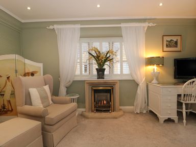 A room with an armchair, a fireplace with flowers, white curtains, a desk with a lamp and a chair at Tree Tops in Broadway