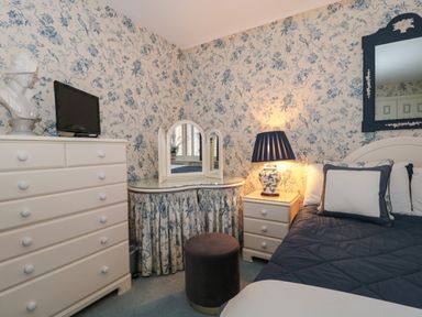A bedroom with blue and white floral wallpaper a dresser with a TV a vanity table with a tri-fold mirror a stool a bedside table with a lamp and a bed with pillows and a quilt at Tree Tops in Broadway