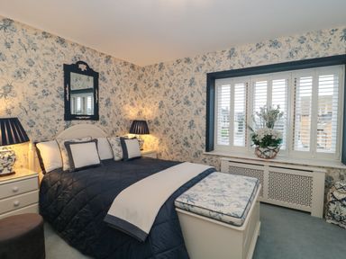 A bedroom with a bed with patterned pillows two bedside tables with lamps a window with shutters and a floral arrangement at Tree Tops in Broadway