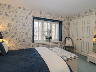 A bedroom with a blue and white bedspread a floral wallpaper window with shutters and a floral chair at Tree Tops in Broadway