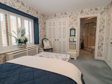 A bedroom with floral wallpaper a bed a floral cushioned bench a chair a tall mirror and an en suite bathroom at Tree Tops in Broadway