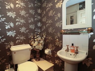 A bathroom with a toilet a pedestal sink a mirror floral wallpaper and a vase with flowers at Tree Tops in Broadway