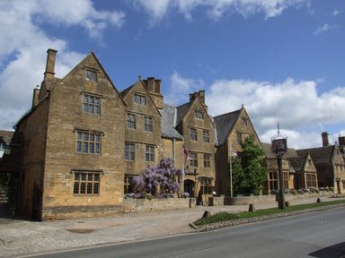 An old stone building with multiple gables and windows along a road with flagpoles and flowering plants