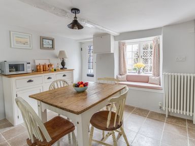 A dining area with a wooden table and chairs a sideboard with a microwave and copper canisters a window seat with pink cushions and flowers at Amy Cottage in West Lulworth