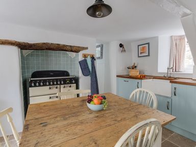 A kitchen with a wooden dining table and chairs a stove with a wooden beam above blue cabinets and a sink at Amy Cottage in West Lulworth