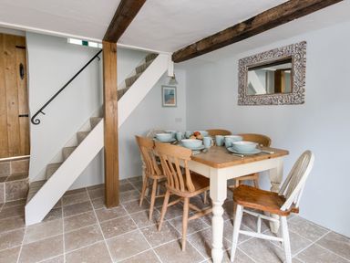 A dining area with a wooden table set with bowls and mugs six chairs a staircase and a decorative mirror on the wall at Amy Cottage in West Lulworth