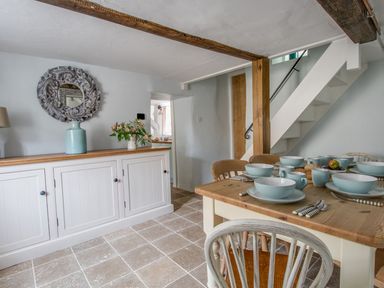 A dining area with a wooden table set with bowls cups and plates next to a white sideboard with a vase and flowers at Amy Cottage in West Lulworth