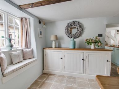A room with a window seat with cushions and a cabinet with a lamp vase flowers and a decorative mirror at Amy Cottage in West Lulworth