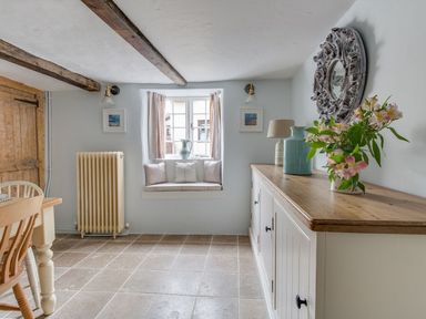 A dining area with wooden chairs and table a sideboard with flowers and vases a window seat with cushions and a radiator at Amy Cottage in West Lulworth