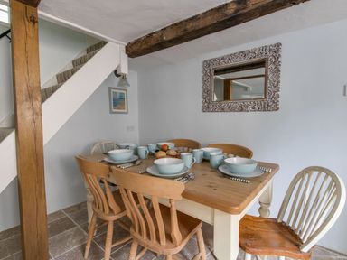 A dining area with a wooden table set with plates cups and cutlery and surrounded by six chairs under a staircase at Amy Cottage in West Lulworth