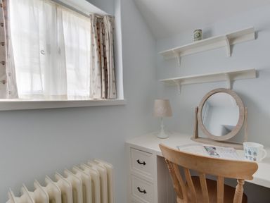 A small room with a wooden chair at a white desk with a mirror lamp and shelves at Amy Cottage in West Lulworth