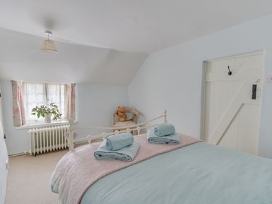 A bedroom with a bed folded towels a window with a vase of flowers a chair with a teddy bear and a door at Amy Cottage in West Lulworth