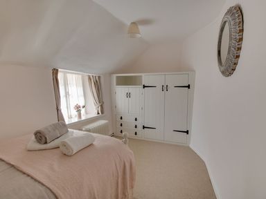 A bedroom with a bed covered by a blanket and towels on it next to a window with curtains and a built in white wardrobe with drawers at Amy Cottage in West Lulworth