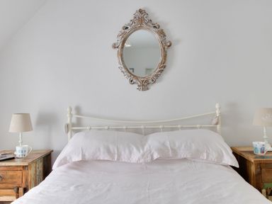 A bedroom with a white metal bed frame white bedding two wooden bedside tables with lamps and mugs and a decorative oval mirror on the wall at Amy Cottage in West Lulworth