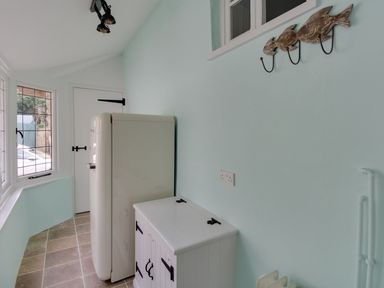 A small utility room with a fridge, white cabinet, fish-shaped wall hooks, and a window at Amy Cottage in West Lulworth