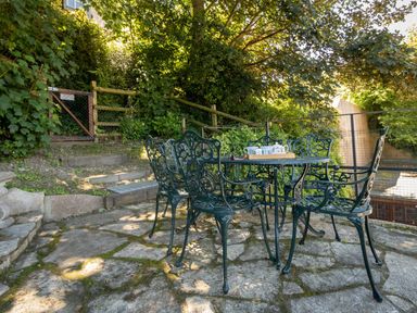 An outdoor patio with a metal table and chairs surrounded by trees and greenery at Amy Cottage in West Lulworth