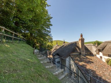 Stone steps with metal railing alongside thatched roof cottages and grassy hill at Amy Cottage in West Lulworth
