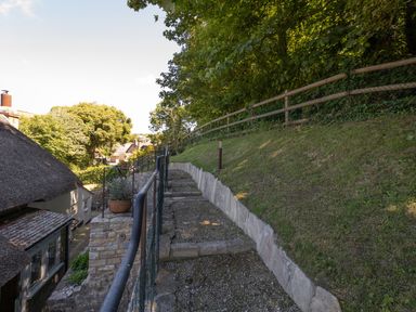An outdoor stone and gravel staircase with metal railing next to a grass slope and thatched roof buildings at Amy Cottage in West Lulworth