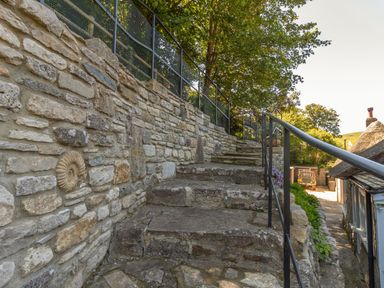 An outdoor stone staircase with metal railing beside a stone wall and trees in the background at Amy Cottage in West Lulworth