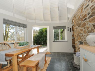 A dining area with wooden table and bench near an open door with stone wall and slate floor at Rose Cottage in Callington