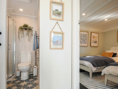 A bathroom with a toilet and shower next to a bedroom with two beds and framed pictures at Rose Cottage in Callington