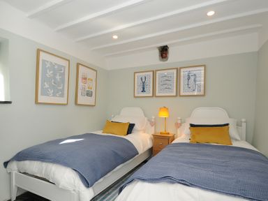 A bedroom with two single beds blue blankets yellow pillows a nightstand with lamp and framed artwork on the walls at Rose Cottage in Callington