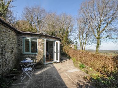 An outdoor patio with a small white table and chairs next to a stone building with open doors and a window at Rose Cottage in Callington