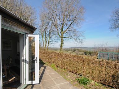 An open glass door leading to a stone patio with a woven wooden fence and trees overlooking the countryside at Rose Cottage in Callington